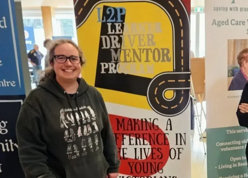 A Bass Coast L2P team member beside the Learner Driver Mentor Program banner at a community event